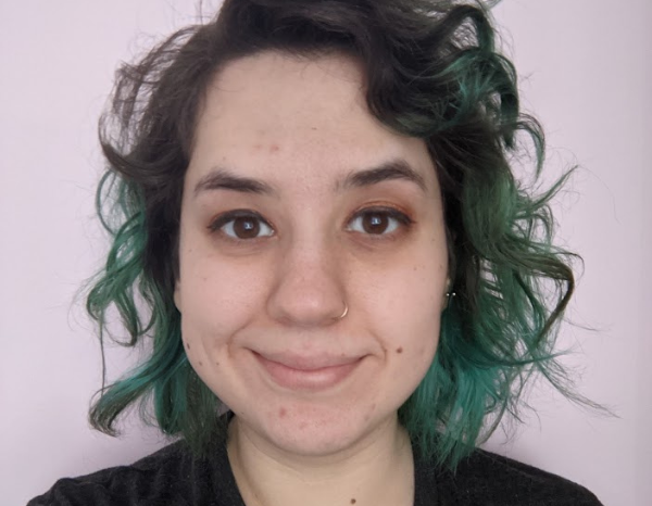 Dasha, a white woman in her 20s with curly dark brown & teal hair, wearing a black T-shirt and smiling, against a white background