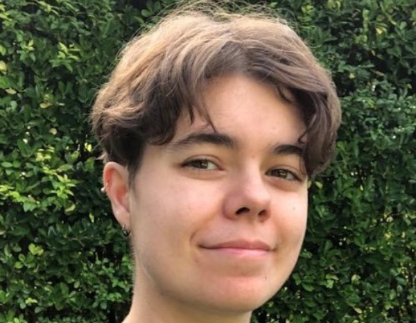 Photograph of Jamie Southwell, a non-binary person - White British, short brown hair In curtains undercut style, brown eyes, wearing a black t-shirt, smiling and looking at the camera with the backdrop of dark green foliage. 