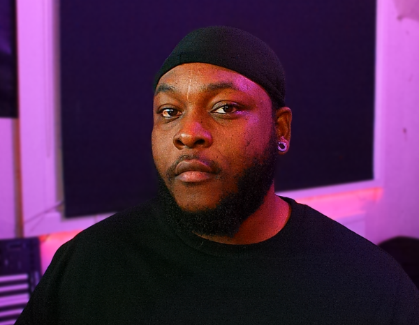 Jason Adenuga, Black Male wearing a black Durag and black crew neck sweater, Sitting in front of a black and pink backdrop.