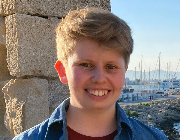 Person with short blonde hair standing in front of a harbour in a blue shirt and red top.