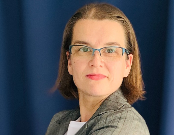 A light-skinned woman with brown jaw length hair. She wears glasses and a gray striped jacket over white T-shirt. The background is blue without any object.