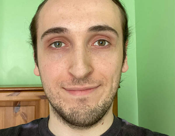 Micky smiling for the camera, in a black t-shirt in front of a green background. 
