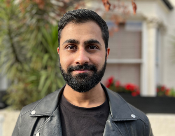 Sheheryar has short black hair, a beard and a moustache, wearing a black T-shirt with a black leather jacket on top and I am standing in front of green trees.