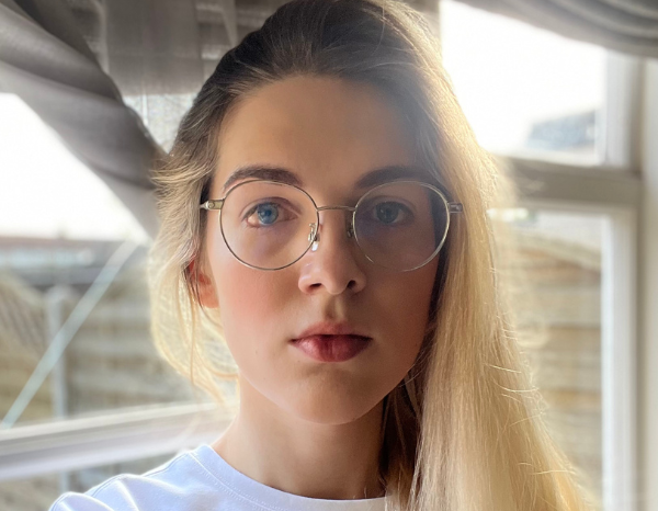 Photo of Skye taken to a blurred backdrop of a London suburb. Skye has blue eyes and long hair fading from brown to blonde, and is wearing a plain white shirt and round glasses.