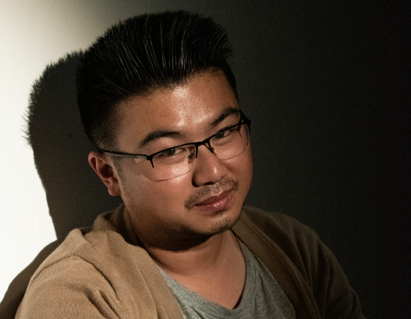 Thanh Tran is sitting up right in a room with a light shining at him, wearing a brown cardigan and grey shirt. The background is being lit in a dark room against a white wall.