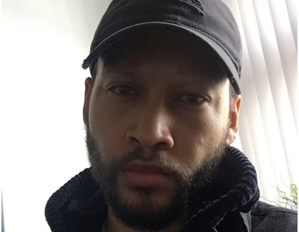 Headshot of a man in a jacket and a cap, standing in an office.