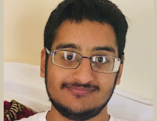 Zain Ali has jet black hair and have a tan skin complexion. He is smiling, wearing a pair of glasses and a white T-shirt against a white background.